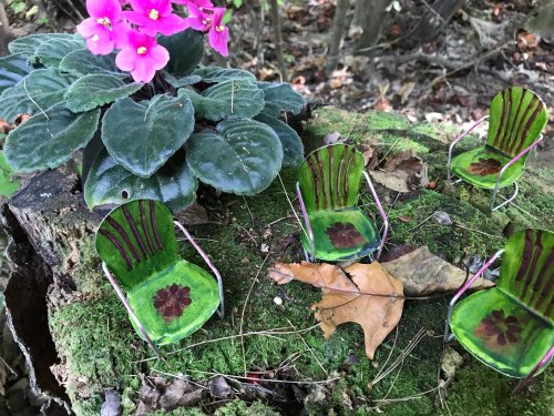 Miniature chairs made of green and pink materials sit on mossy ground near a flowering plant.