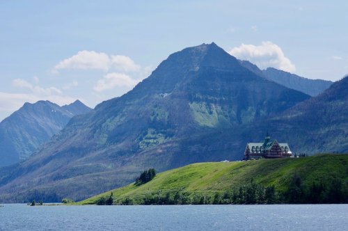 A scenic view of a green hillside with a large building, surrounded by mountains and a calm lake under a blue sky.