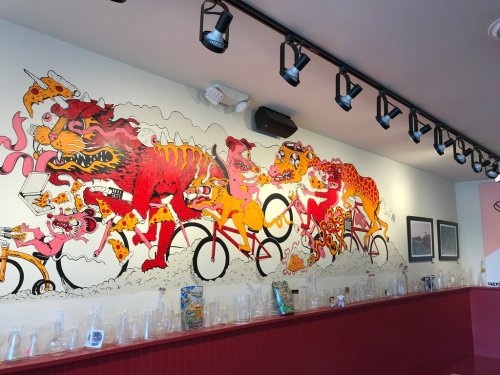 Colorful mural featuring cartoon animals on bicycles, surrounded by pizza, with a glass display in the foreground.