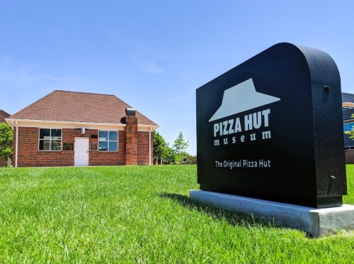 A sign for the Pizza Hut Museum in front of a small brick building on a grassy area under a clear blue sky.