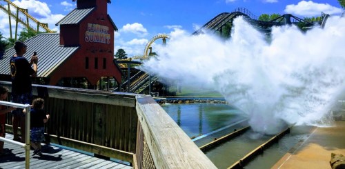 A water ride splashes down near a wooden deck, with roller coasters in the background and people watching.