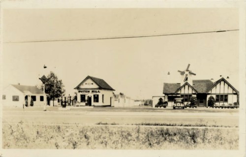 Historic black-and-white photo of Dutch Mill buildings along a road, featuring a windmill and vintage cars.