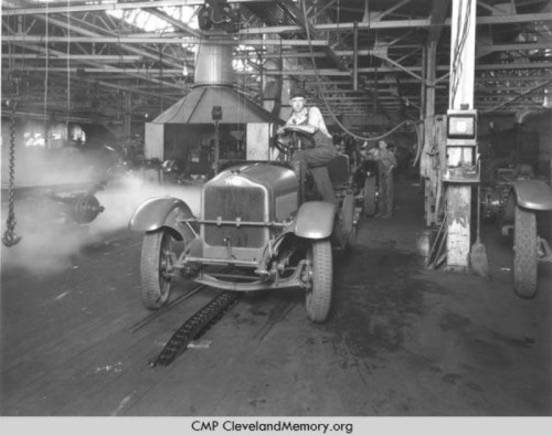 A vintage factory scene with a man driving an early 20th-century vehicle amidst machinery and smoke.