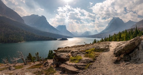 A panoramic view of a serene lake surrounded by mountains, with sunlight breaking through clouds and reflecting on the water.