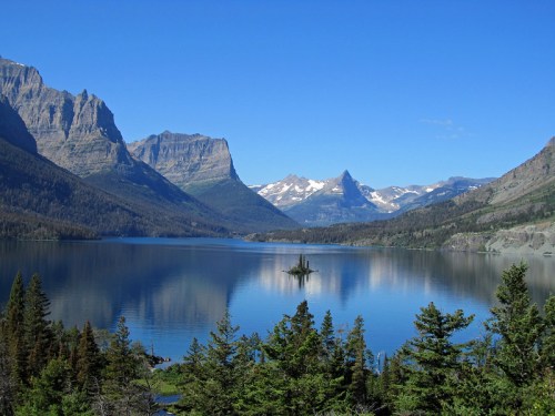 A serene lake surrounded by mountains, with a small island in the center and clear blue skies above.