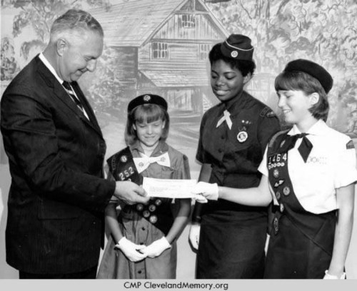 A man presents a check to three Girl Scouts in uniforms, smiling in front of a painted backdrop.