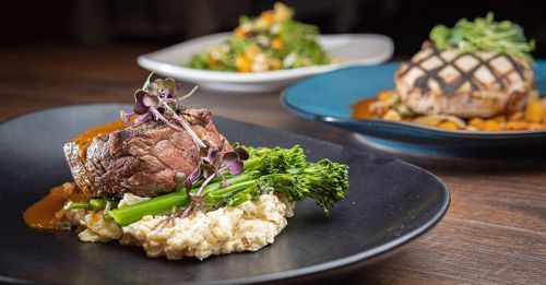 A plated dish featuring a beef filet on creamy mashed potatoes, garnished with broccolini and microgreens, with other dishes in the background.
