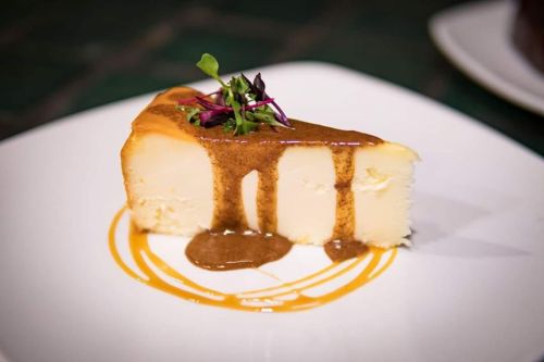 A slice of cheesecake drizzled with caramel sauce and garnished with microgreens on a white plate.