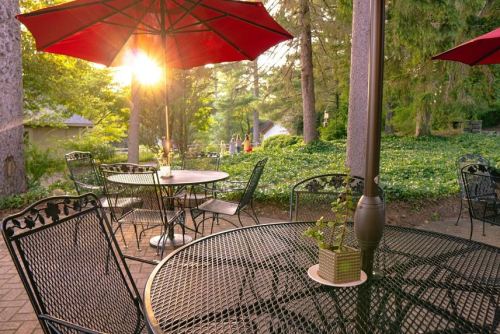 A sunlit outdoor café with metal tables and red umbrellas, surrounded by greenery and trees.