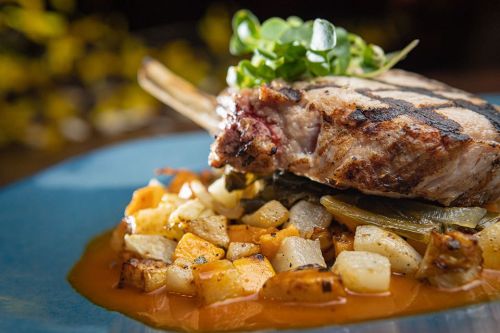 Grilled pork chop on a bed of roasted vegetables, garnished with microgreens, served on a blue plate.