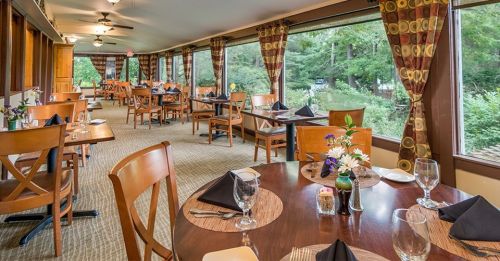 A cozy restaurant interior with wooden tables, chairs, and large windows overlooking greenery.