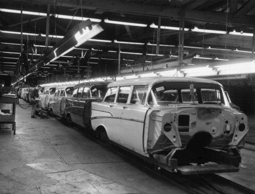 Black and white image of a car assembly line with several unfinished vehicles in a factory setting.