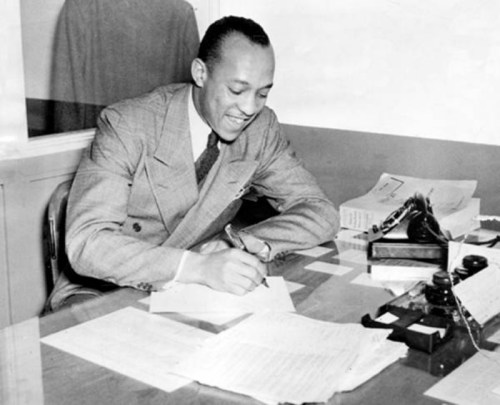 A smiling man in a suit sits at a desk, writing on paper with documents and a typewriter nearby.
