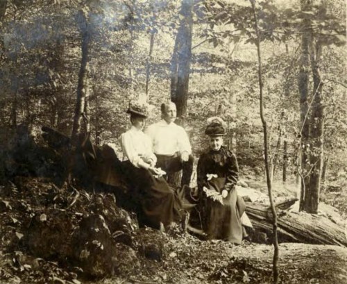 Three people in vintage clothing sit on a log in a wooded area, surrounded by trees. The photo has a sepia tone.
