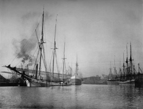 Black and white image of several sailing ships docked in a harbor, with smoke rising in the background.