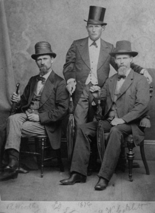 Three men in formal attire, including top hats and suits, pose together with umbrellas in a vintage studio setting.