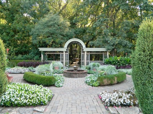 A serene garden with lush greenery, colorful flowers, and a central fountain surrounded by decorative arches.