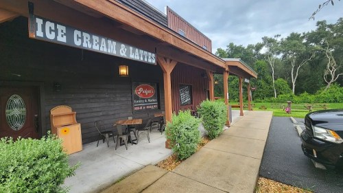 Exterior of an ice cream shop with outdoor seating, surrounded by greenery and a cloudy sky.