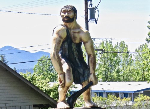 A large statue of a muscular man holding a club, set against a backdrop of trees and mountains.