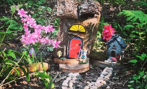 A whimsical fairy house made from wood, surrounded by flowers and a stone path in a lush green forest setting.
