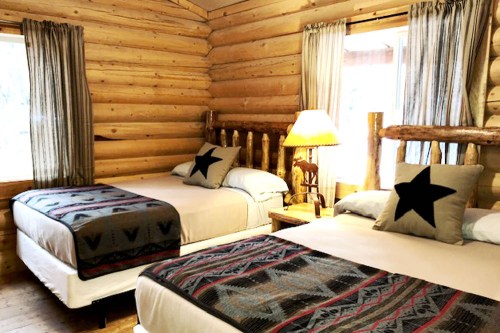 Cozy log cabin bedroom with two beds, star-patterned pillows, and rustic decor, featuring wooden walls and natural light.