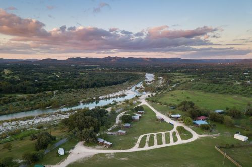 Aerial view of a winding river surrounded by green fields and hills under a colorful sky with scattered clouds.