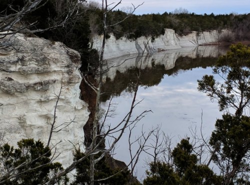 A serene landscape featuring white cliffs reflected in calm water, surrounded by trees and a clear sky.