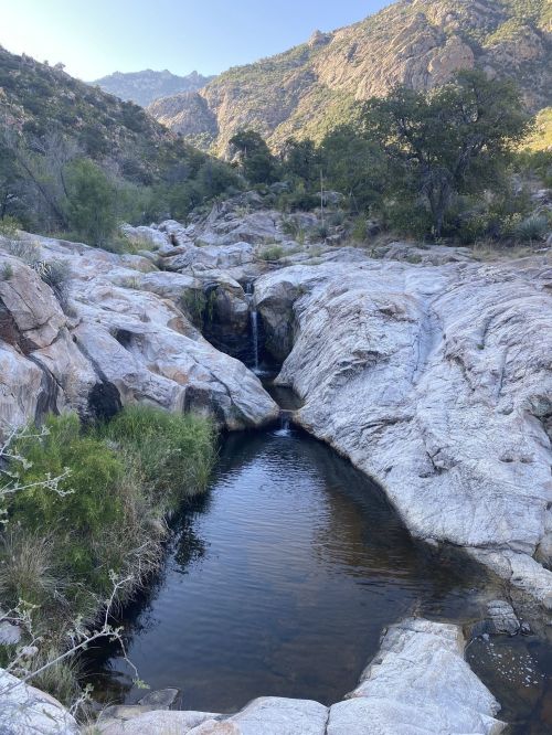 A serene rocky landscape with a small waterfall flowing into a clear pool, surrounded by greenery and mountains.