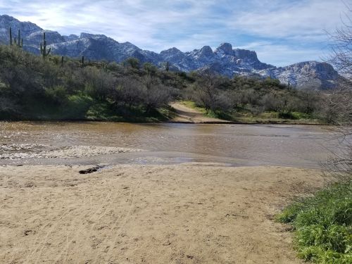A sandy riverbank with a calm waterway, surrounded by mountains and sparse vegetation under a clear blue sky.
