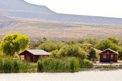 A serene landscape featuring two wooden cabins by a lake, surrounded by lush greenery and mountains in the background.