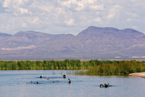 People swimming in a calm lake surrounded by lush greenery and mountains under a partly cloudy sky.