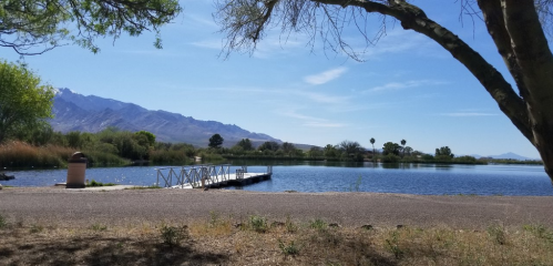 A serene lake scene with a wooden dock, surrounded by trees and mountains under a clear blue sky.