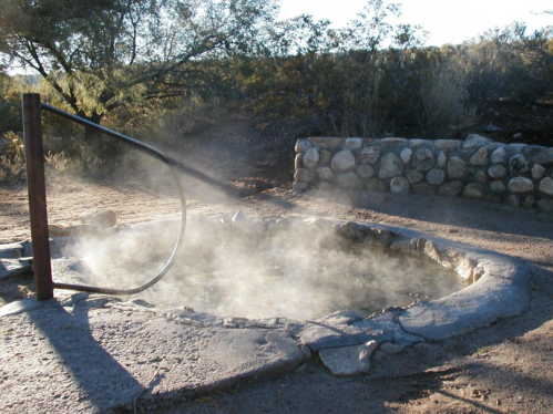 A steaming hot spring surrounded by rocky terrain and sparse vegetation under a clear sky.