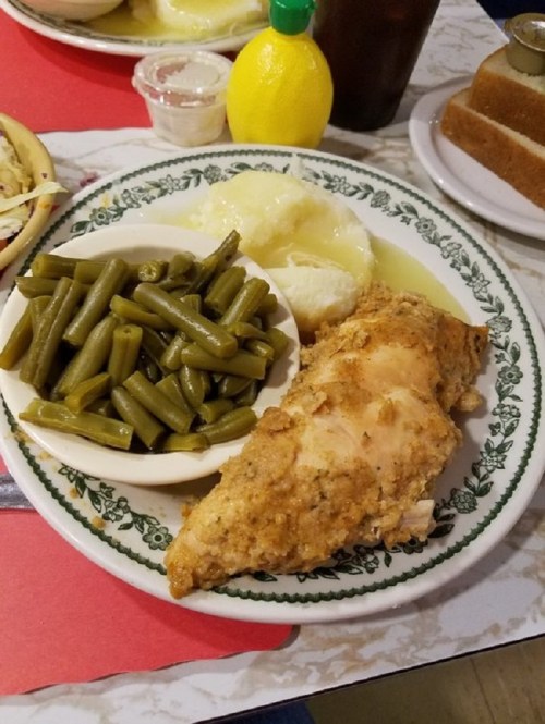 A plate of fried chicken with green beans and mashed potatoes, served with a lemon wedge and a drink in the background.