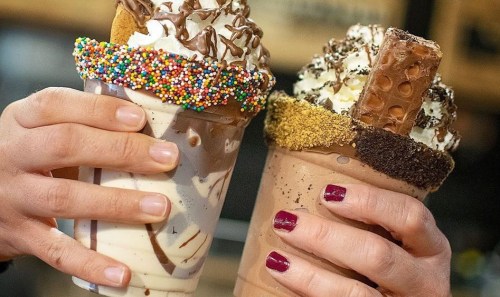 Two hands hold large milkshakes topped with whipped cream, chocolate, sprinkles, and candy bars.
