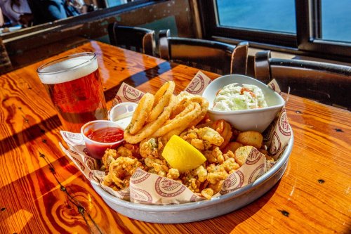 A platter of fried seafood with onion rings, coleslaw, dipping sauces, and a glass of beer on a wooden table.