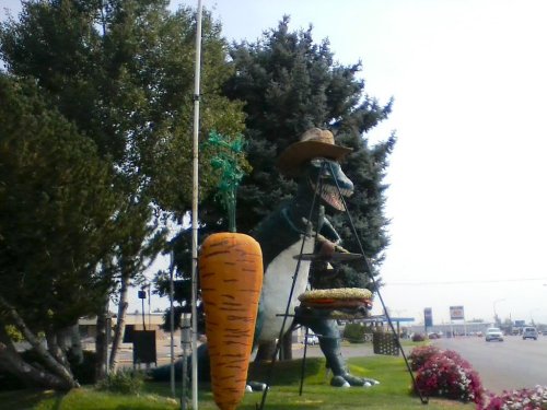 A large dinosaur statue wearing a sombrero, holding a burger, next to a giant carrot and surrounded by trees and flowers.