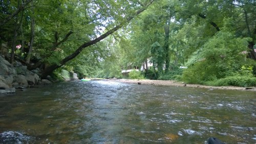 A serene river flows through a lush green landscape, with trees lining the banks and a hint of a house in the background.