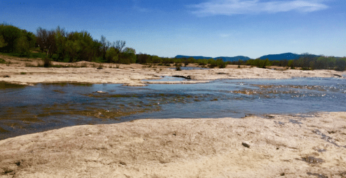 A serene river flows through a sandy landscape, surrounded by trees and distant mountains under a clear blue sky.