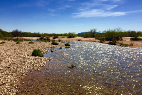 A serene river flows through a rocky landscape, surrounded by sparse vegetation and distant mountains under a clear blue sky.
