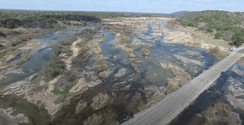 Aerial view of a winding riverbed with sparse vegetation and a road crossing through a dry landscape.