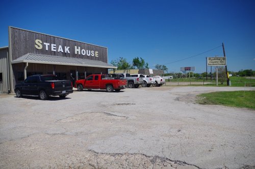 A steakhouse with a wooden exterior and several parked trucks in front, set against a clear blue sky.