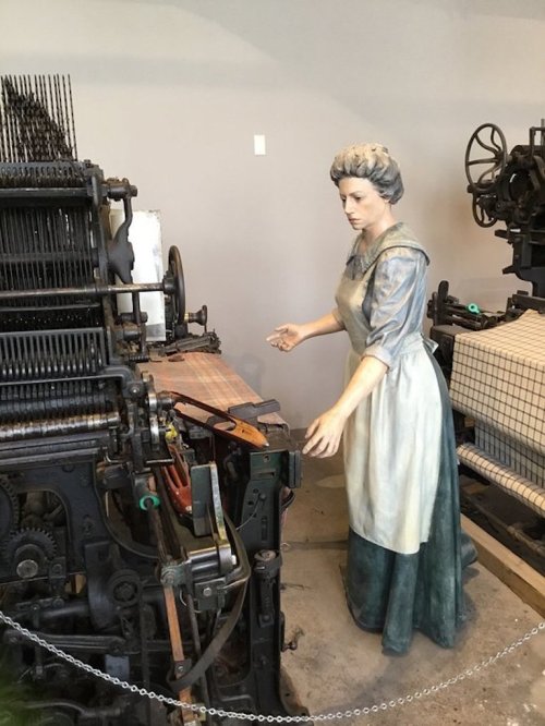 A life-sized statue of a woman in period clothing operating an old printing press in a museum setting.