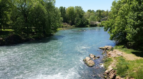 A serene river flows through a lush green landscape, with trees lining the banks and rocky outcrops along the shore.