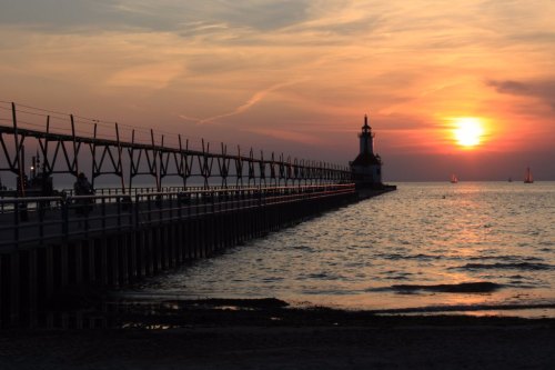 A serene sunset over a pier with a lighthouse, boats in the distance, and calm waters reflecting the warm colors.