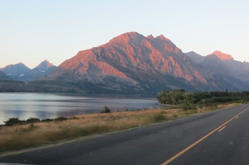 A scenic view of a mountain bathed in sunset light, beside a calm lake and a winding road.