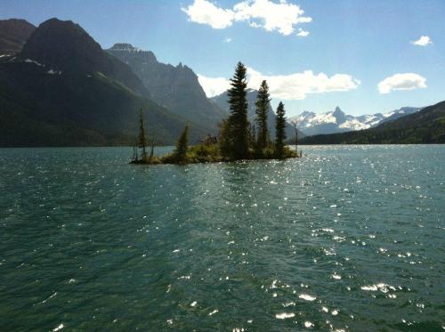 A serene lake scene featuring a small island with trees, surrounded by mountains and a clear blue sky.