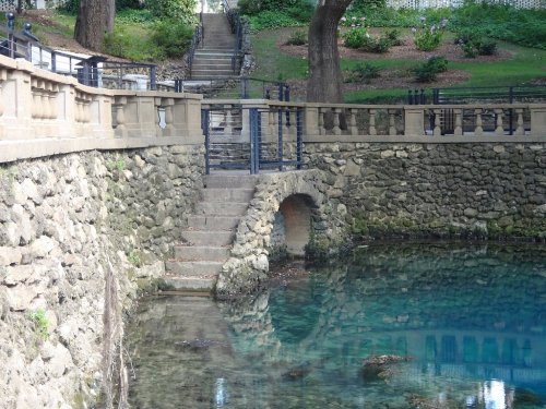 A stone bridge leads to a clear pond, surrounded by trees and a staircase in a serene park setting.