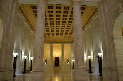 Interior of a grand hall with marble columns, ornate ceiling, and soft lighting from wall-mounted lamps.