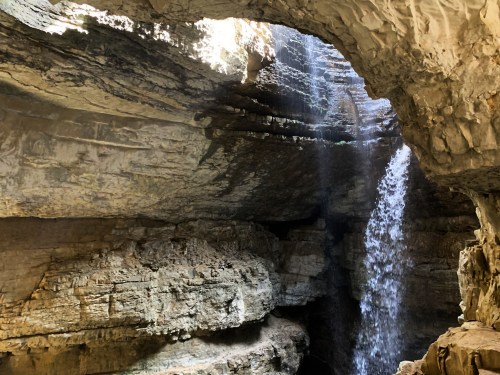 A waterfall cascades down rocky cliffs inside a cave, illuminated by sunlight filtering through the opening above.